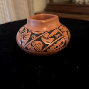 Może przedstawiać: Mały, ręcznie malowany wazon ceramiczny z geometrycznym wzorem w kolorze czarnym na tle terakoty. Wazon ma zaokrąglone ciało i kwadratowy otwór.