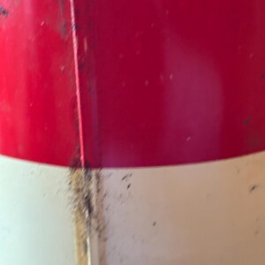 May include: Close-up of a red and white painted metal object. The top portion is red, and the bottom is white. A vertical seam runs down the center, with some discoloration and debris.