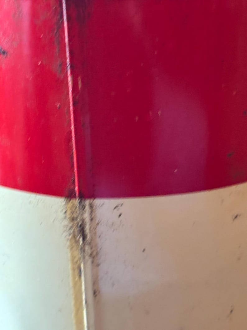 May include: Close-up of a red and white painted metal object. The top portion is red, and the bottom is white. A vertical seam runs down the center, with some discoloration and debris.