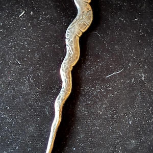 Peut inclure: Un ouvre-lettre argenté au design incurvé et allongé. Le haut présente un relief détaillé d'une figure classique. La poignée a un motif texturé et ondulé, et l'extrémité a un crochet incurvé. L'ouvre-lettre est probablement en métal.