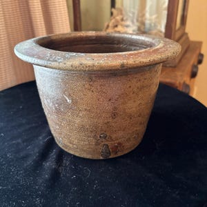 Könnte beinhalten: Ein brauner Keramiktopf im antiken Stil mit einem breiten, abgerundeten Rand. Der Topf hat eine zylindrische Form und eine strukturierte Oberfläche mit subtilen Farbvariationen. Er scheint gealtert zu sein, mit kleinen Unvollkommenheiten, die zu seinem Vintage-Appeal beitragen. Der Topf ist wahrscheinlich für dekorative oder funktionale Zwecke.