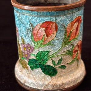Pode incluir: Uma pequena caixa redonda de cobre com um design de esmalte azul e branco. A caixa é decorada com flores rosa, vermelhas e verdes.