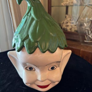 May include: A ceramic jar shaped like an elf head with a glossy white face, large pointed ears, blue eyes, and a red mouth. The lid is a removable green hat with a textured, leaf-like design that curves at the top, completing the elf character.