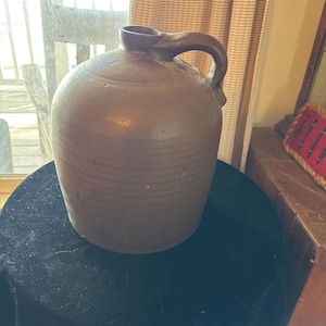 May include: A brown stoneware jug with a rounded body and a handle. The jug has a dark brown color and a textured surface. It is sitting on a black surface. The jug is an antique and is likely used for storing liquids.