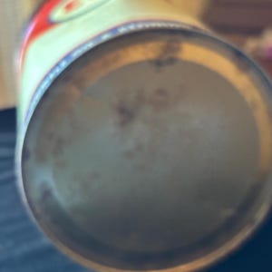 May include: A vintage metal can with a faded design. The can's top features red and white accents with a blue border. The bottom of the can shows signs of age and wear, with a slightly rusted appearance. The can is cylindrical.