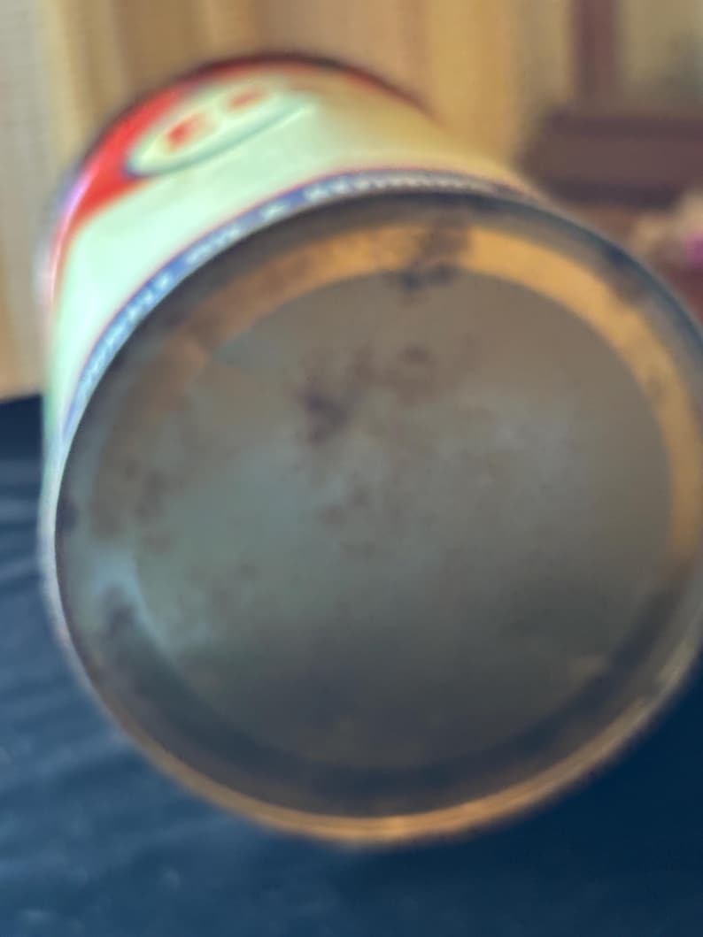May include: A vintage metal can with a faded design. The can's top features red and white accents with a blue border. The bottom of the can shows signs of age and wear, with a slightly rusted appearance. The can is cylindrical.