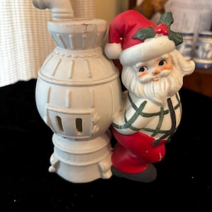 May include: A vintage ceramic Christmas figurine featuring Santa Claus and a white stove. Santa is wearing a red hat and suit with a green and black plaid shirt. The stove is white with a curved chimney.