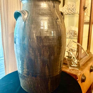 May include: A large, antique brown stoneware crock with two side handles. The crock has a wide mouth and a dark, glossy finish. The surface shows variations in color, with lighter and darker brown tones. This decorative piece is suitable for home decor.