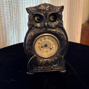 May include: A dark metallic owl-shaped clock featuring textured feather details and two prominent green eyes. The circular clock face is set into the centre of the owl's body, displaying black numerals from one to twelve and thin black hands.