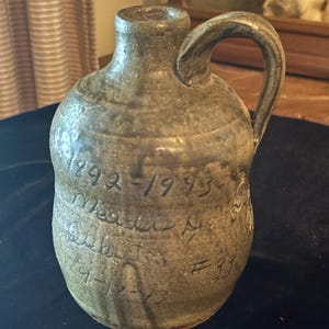 May include: A handcrafted, stoneware jug with a handle, featuring a mottled, earthy glaze. The jug has handwritten inscriptions including dates and the number "39". The jug is a rustic, vintage piece.