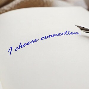 Op de afbeelding: Een open notitieboek met de handgeschreven zin "I choose connection" in blauwe inkt. Een zilveren en houten vulpen ligt op de pagina. Een neutraal gekleurde gebreide deken en een witte mok staan op de achtergrond.