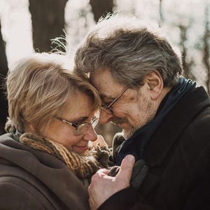May include: A tender moment captured between an older couple. The woman, with short blonde hair and glasses, leans her head against the man's. He has gray hair, glasses, and a beard. They are embracing, with the man's hand gently touching the woman's.
