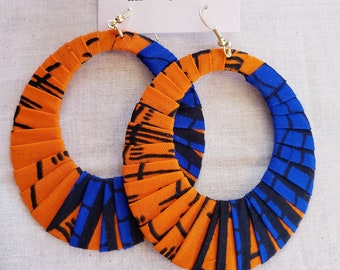 May include: A pair of large hoop earrings. The earrings are made of orange fabric with black and blue geometric patterns. The earrings have silver-colored hooks and are displayed on a white background. The text "UWAK DESIGNS" is visible.