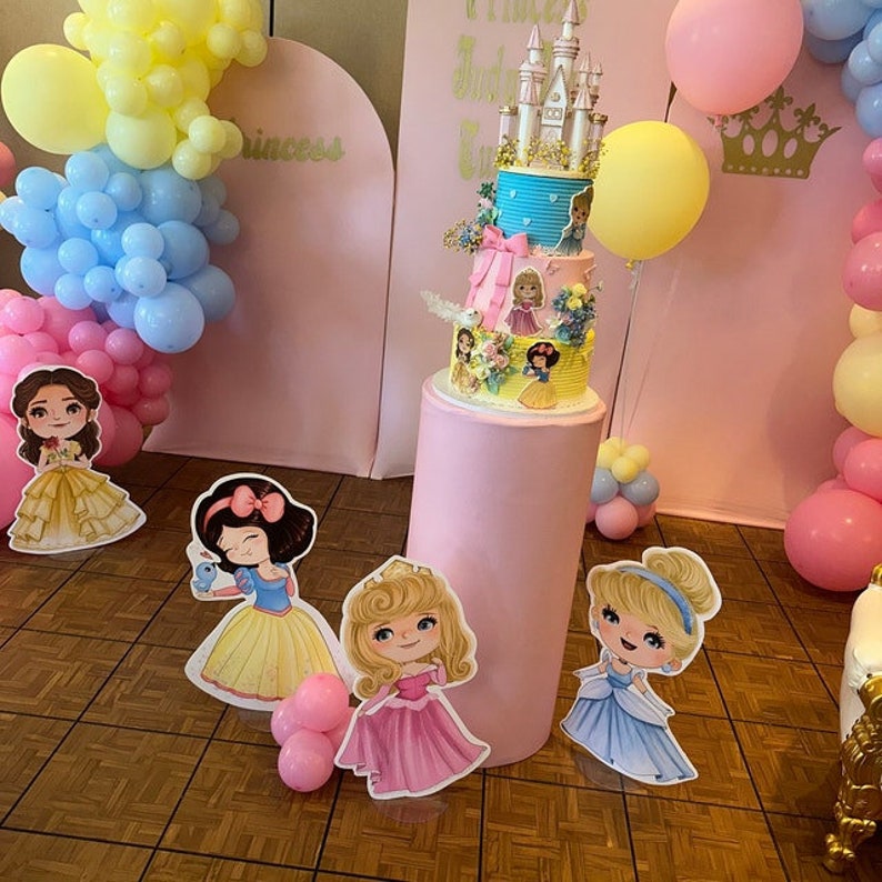 May include: A pink and blue princess-themed cake with a castle on top. The cake is surrounded by balloons and princess cutouts. The cake is on a pink pedestal.