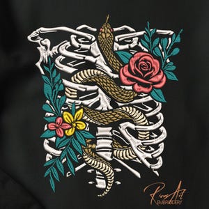 Könnte beinhalten: Gesticktes Design mit einer Schlange, die sich um einen Brustkorb windet, mit einer roten Rose und bunten Blumen. Die Schlange ist braun und goldfarben, die Blumen sind rosa und gelb, und die Blätter sind türkis. Das Design befindet sich auf schwarzem Hintergrund.
