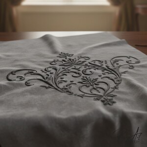 May include: A gray textile with an embroidered black floral design. The design is centered and features intricate swirls, leaves, and flower-like shapes. The fabric is draped, and the embroidery has a raised texture, creating a three-dimensional effect.