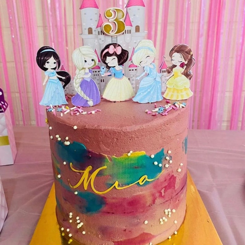 May include: A pink and white cake with a rainbow swirl design and the name "Mia" written in gold. The cake is decorated with princess figurines and a castle, and a gold number "3" candle.