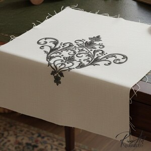 May include: Cream-colored linen table runner with a black embroidered floral design in the corner. The design features swirling lines and leaf shapes. The runner is displayed on a dark wooden desk.
