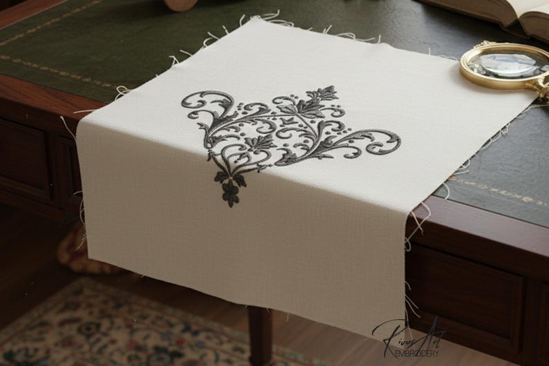 May include: Cream-colored linen table runner with a black embroidered floral design in the corner. The design features swirling lines and leaf shapes. The runner is displayed on a dark wooden desk.