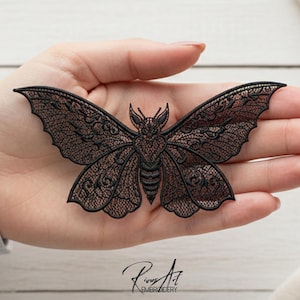 May include: Black embroidered moth applique with intricate lace-like detailing. The moth has a detailed body and wings, with a delicate pattern. The image includes the text "Rivus Art Embroidery".