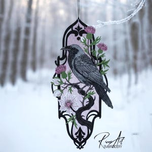 Könnte beinhalten: Gesticktes Kunstwerk mit einer Krähe auf einem Ast mit rosa Disteln und weißen Blumen, eingebettet in einen dekorativen schwarzen und rosa Rahmen. Das Werk wird von einer weißen Schnur gehalten, mit einem verschneiten Wald im Hintergrund.