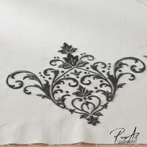 May include: Close-up of a detailed, dark gray embroidered floral design on a white fabric. The embroidery features a symmetrical pattern of leaves, swirls, and dots, with a heart-shaped element at the center. The design is likely for home decor or textile crafts.