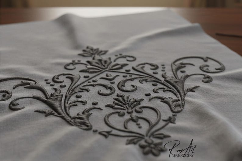 May include: Close-up of a gray fabric with an embroidered dark gray floral design. The embroidery features a symmetrical pattern of swirling leaves, flowers, and scrollwork. The fabric is slightly wrinkled, and the embroidery has a raised texture.
