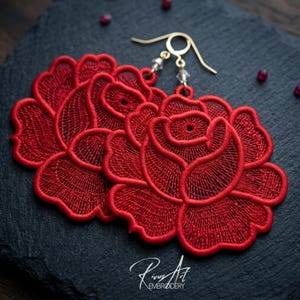 May include: Red embroidered rose earrings with gold-colored hooks and clear bead accents. The earrings are displayed on a dark gray textured surface, with small red beads scattered around them. The earrings are a floral design.