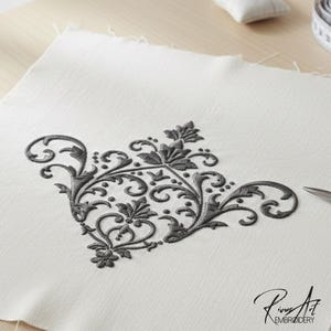 May include: A detailed, dark gray embroidered floral design on a square of white fabric. The design features swirling patterns and leaf motifs. Scissors and a measuring tape are visible, suggesting a craft or sewing project. The image includes the text "Rinus Art Embroidery".