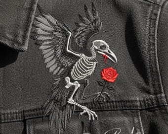 May include: Dark grey denim jacket with an embroidered design of a skeletal raven holding a red rose. The raven has outstretched wings and a beak with a drop of red. The jacket has a button closure and a small logo at the bottom.