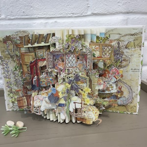 May include: A detailed pop-up book featuring a whimsical scene with illustrated animals in a cozy, detailed setting. The book's pages are open, revealing a three-dimensional diorama with furniture, flowers, and characters. The color palette includes soft greens, browns, and purples.