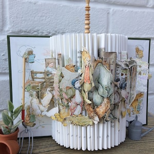 May include: A book sculpture featuring characters from Beatrix Potter's children's book, The Tale of Peter Rabbit. The sculpture is made from pages of a book that have been folded and glued together to create a three-dimensional scene. The characters include Peter Rabbit, Mr. McGregor, and the ducks. The sculpture is topped with a wooden finial.