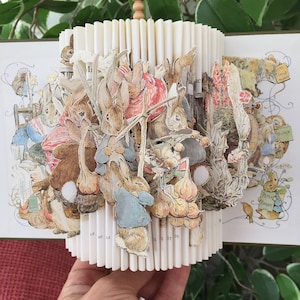 Peut inclure: Une sculpture d'art de livre représentant les personnages de Peter Rabbit du livre pour enfants de Beatrix Potter. La sculpture est faite de pages pliées du livre, avec des images découpées de Peter Rabbit et d'autres personnages. La sculpture est tenue dans une main, avec un fond de feuilles vertes.
