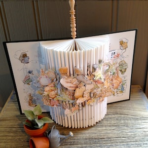 May include: A book art sculpture featuring a 3D pop-up design of a group of rabbits and other woodland creatures. The book is open and fanned out, creating a circular shape. The sculpture is made from pages of a book that have been cut and folded. The sculpture is decorated with illustrations of rabbits, squirrels, and other woodland creatures. The sculpture is mounted on a wooden base.