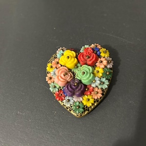 Puede incluir: Broche en forma de corazón con un marco dorado, lleno de flores coloridas. El broche presenta una variedad de tipos de flores en tonos de amarillo, rojo, rosa, verde y morado. Un encantador accesorio floral.