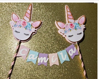 May include: Two unicorn cake toppers featuring floral crowns, closed eyes, and gold glitter horns, connected by a bunting banner with pastel pink, blue, and purple pennants spelling the name Camila in white letters, attached to striped gold straws.