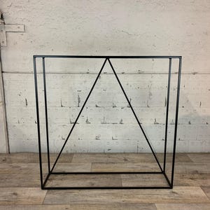 May include: Black metal geometric stand with a square base and a triangular top. The stand is made of thin metal and has a simple, modern design.