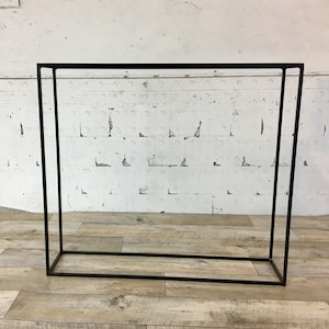 May include: A black metal rectangular frame standing on a wooden floor. The frame is empty and has a simple design.