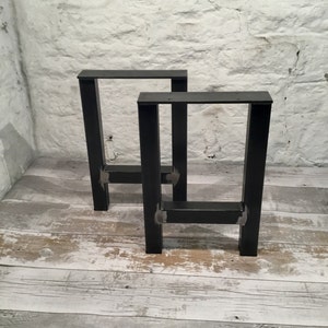 May include: Two black metal table legs with a square design. The legs are made of thick metal and have a simple, industrial look.