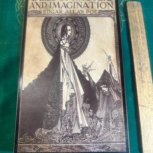 May include: A framed print of "Tales of Mystery and Imagination" by Edgar Allan Poe. The artwork features a detailed black and white illustration of a figure with a halo, and other figures in a gothic style. A wooden ruler is next to the frame.