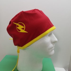 May include: A red Flash-themed scrub cap with a yellow lightning bolt logo on the side. The cap has a yellow trim and yellow ties. The cap is displayed on a white mannequin head.