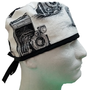 May include: A white scrub hat with a black trim and a black tie. The hat has a pattern of vintage cameras in black and white.