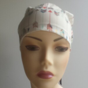 May include: White surgical cap with a colorful arrow print design. The cap has a tie at the back.