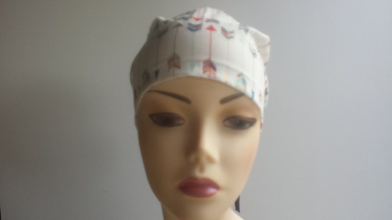 May include: White surgical cap with a colorful arrow print design. The cap has a tie at the back.