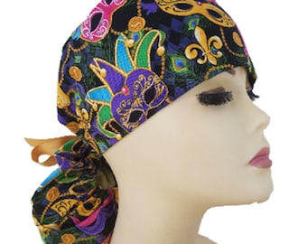 May include: A black scrub cap with a colorful Mardi Gras mask pattern. The cap features gold, purple, green, and pink mask designs, fleur-de-lis, and peacock feathers. It has a ribbon tie in the back.