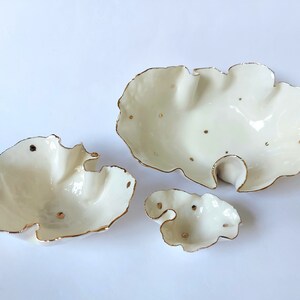 May include: Three white ceramic bowls with irregular, organic shapes and wavy, uneven edges. Each bowl features a glossy finish with metallic gold trim along the rims and small, scattered gold dots across the interior surfaces of the vessels.