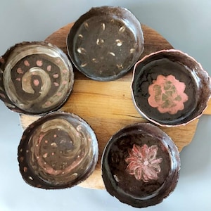 May include: Five handmade ceramic bowls with dark brown exteriors and decorative interiors. Each bowl features unique pink and beige painted designs, including spirals and floral patterns. The bowls are arranged on a wooden chopping board.
