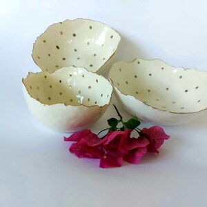 May include: Three off-white ceramic bowls with irregular, wavy rims and gold-painted edges. The interior and exterior surfaces are decorated with small, scattered gold polka dots. A cluster of bright pink bougainvillea flowers sits in front.
