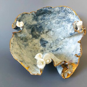 May include: A white ceramic bowl with a blue and white marbled design and gold accents. The bowl has a ruffled edge and three white ceramic flowers on the rim.
