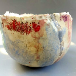 Könnte beinhalten: Eine handgefertigte Keramikschale mit einem rauen, strukturierten Rand. Die Schale zeigt eine Mischung aus Farben, darunter Rot, Blau und Creme, mit einem gesprenkelten, künstlerischen Design. Die Außenseite der Schale hat eine einzigartige, organische Form.